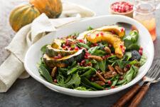 A white bowl filled with roasted acorn squash, spinach, and pomegranates with Acorn squash in the background.