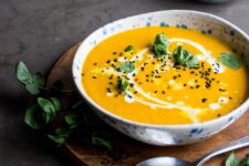 A bowl of turmeric soup with coconut milk drizzled in it. The bowl is on a wooden platter with a smaller bowl of turmeric soup and some herbs.