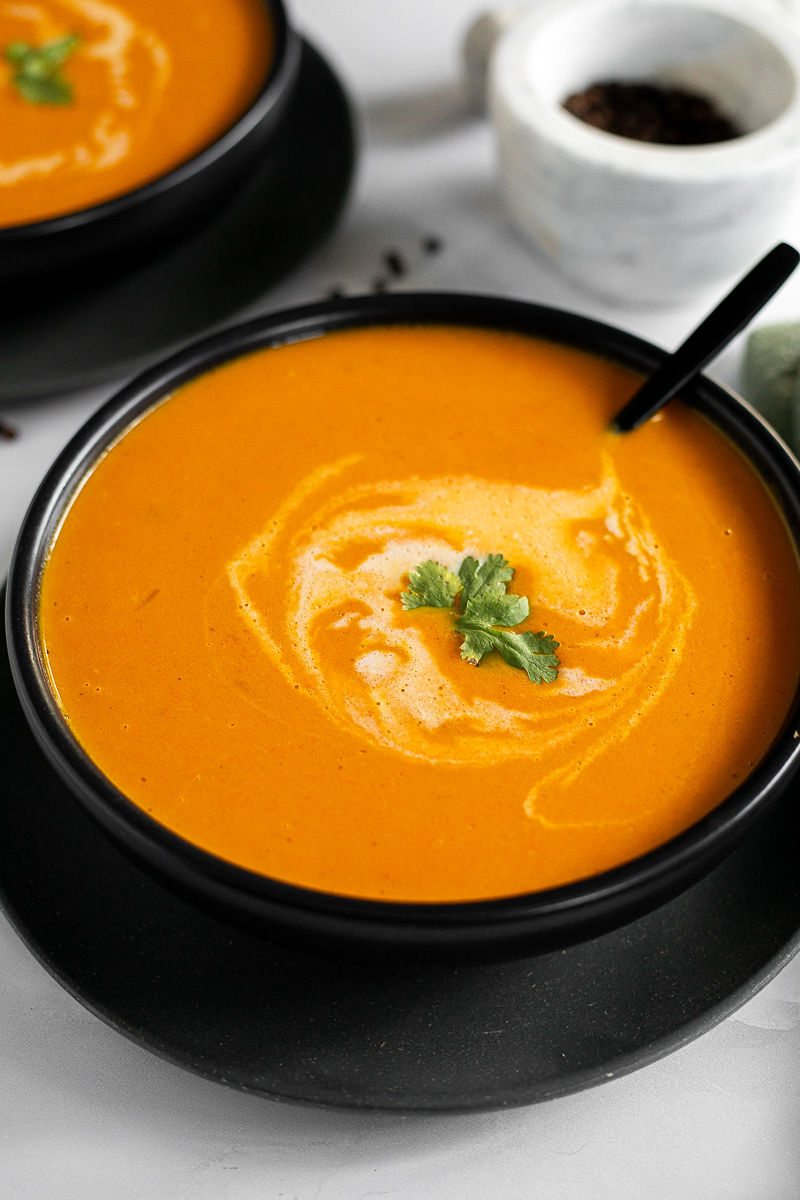 A black soup bowl with coconut carrot soup, garnished with cilantro, and a spoon. 