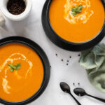 Two bowls of coconut carrot soup with two spoons and a green napkin.