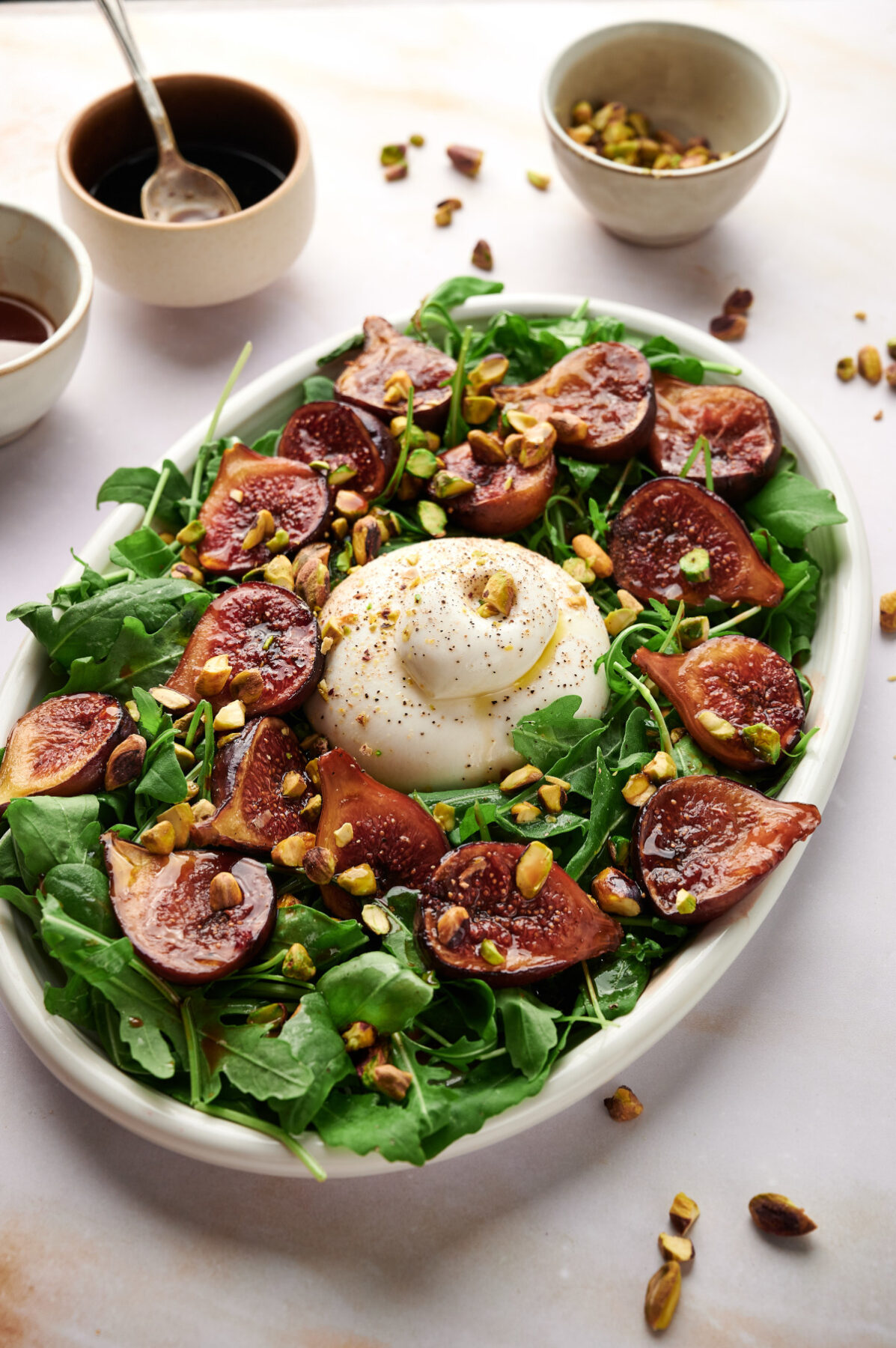 Fig & Arugula Salad with Burrata on a white plate with bowls of condiments and garnishes in the background.