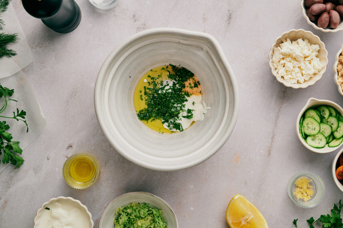 Ingredients for a Greek chicken marinade in a white bowl with smaller bowls of various ingredients.