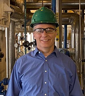 A man wearing a green hard hat, safety glasses and blue collared shirt, standing in front of mechanical equipment