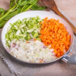 A skillet with diced celery, carrots, and onions. Celery stalks and carrots are in the background.