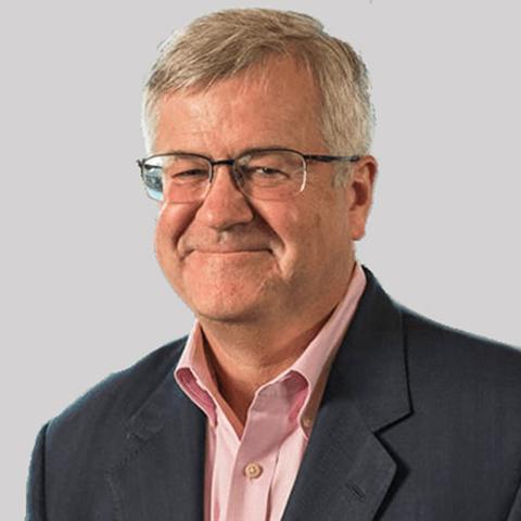 A man with grey hair, glasses, wearing a pink shirt and dark jacket