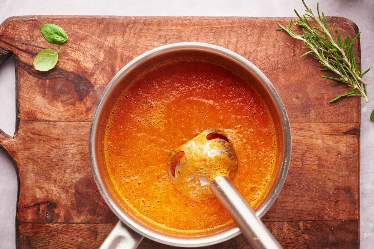 An immersion blender in a pot mixing up some tomato soup on top of a wooden cutting board.