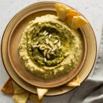 close up image of a pesto hummus in a round plate with pita chips