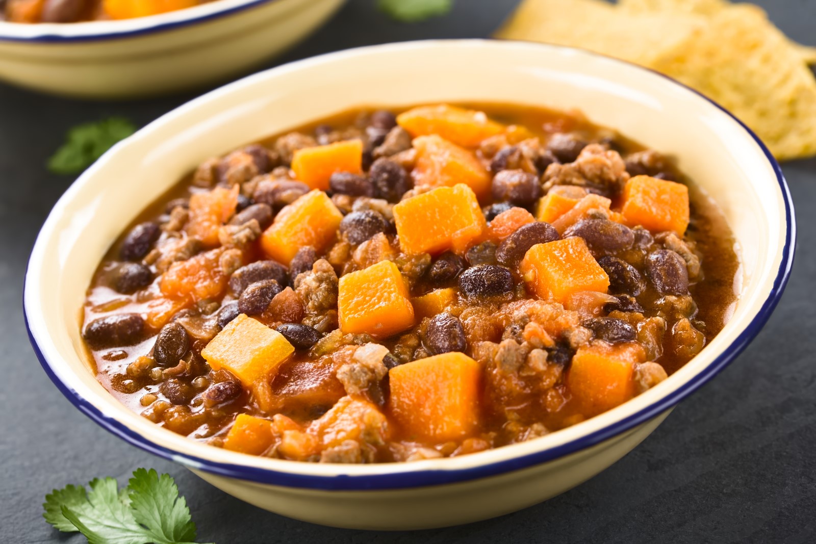 A white bowl with a blue rim filled with pumpkin chili with tortilla chips in the background.