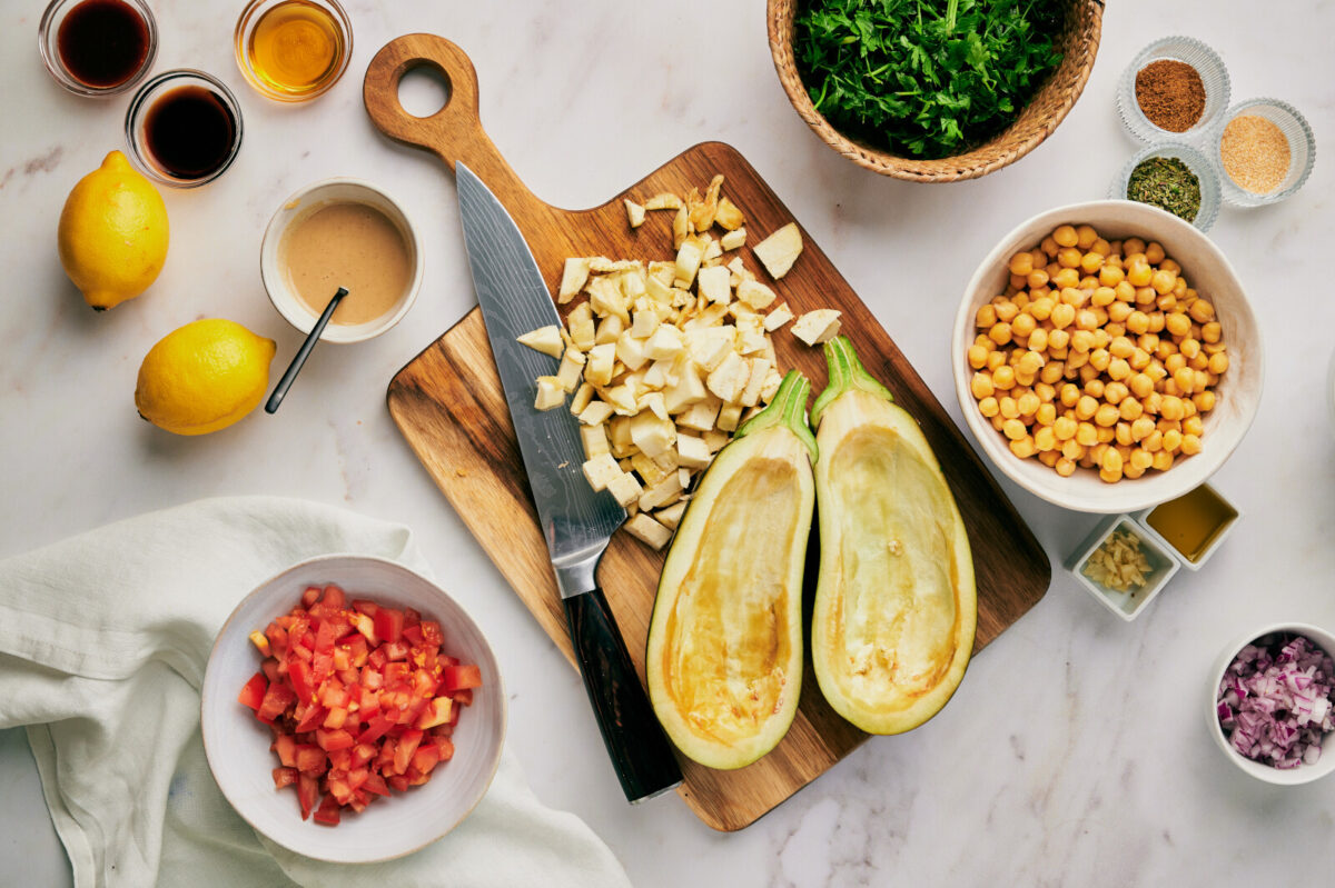 Two egglants with seeds scooped out on a cutting board with more chopped eggplant and a knife surrounded by other ingredients.