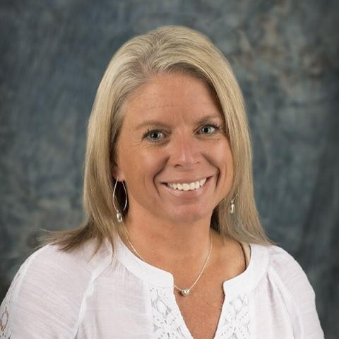 A woman with shoulder-length blonde hair, white shirt, in front of a grey background.