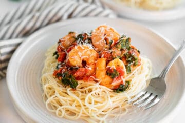 Spicy shrimp and kale served over angel hair pasta on a white plate.