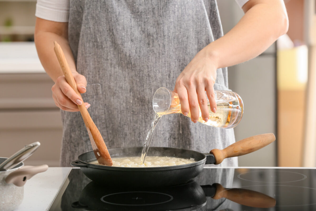 Woman cooking risotto in kitchen. Pouring dry white wine into the Arborio rice mixture.