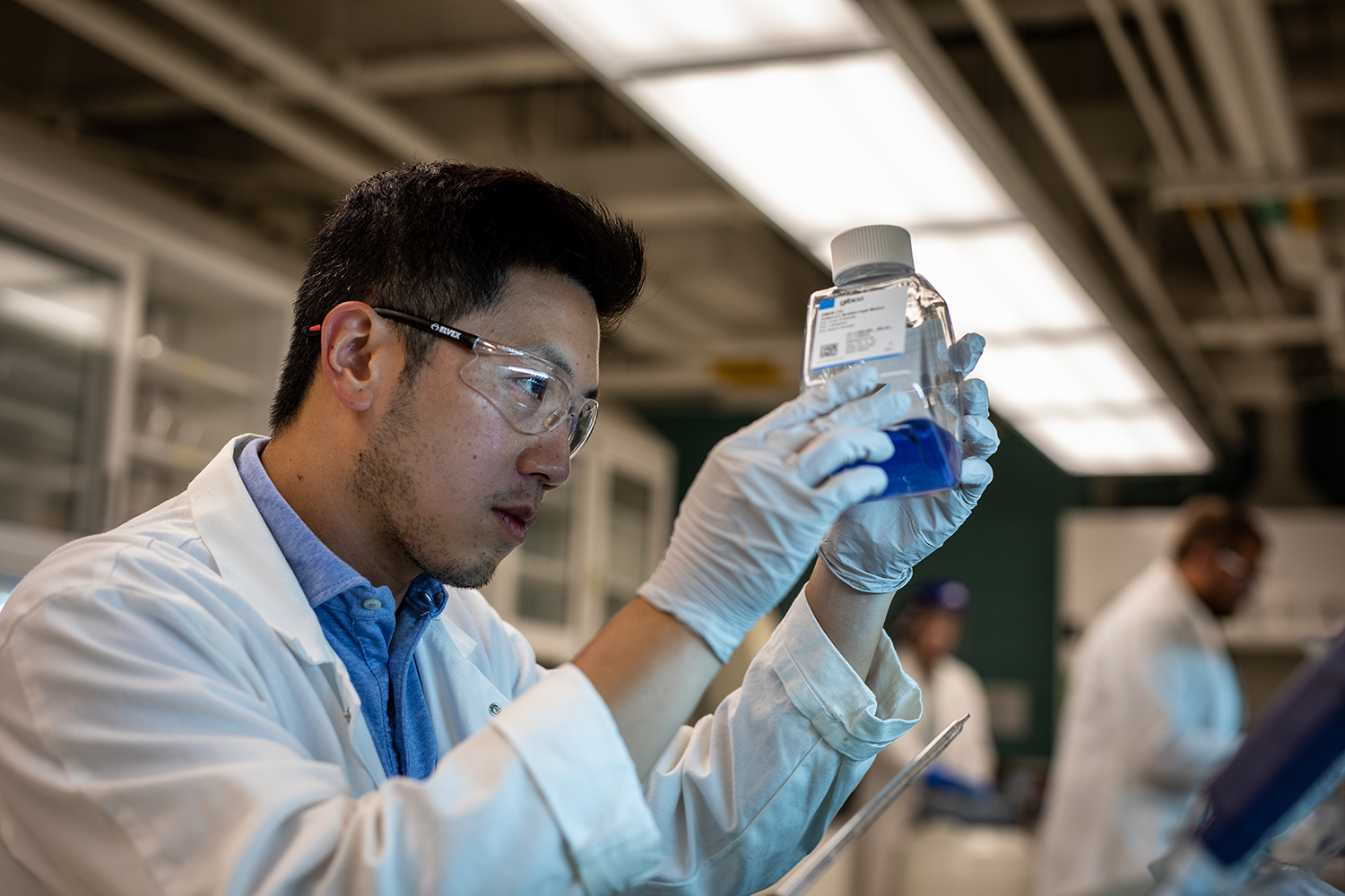 A person wearing a lab coat, gloves, and safety glasses, looking into a glass bottle with blue liquid. 