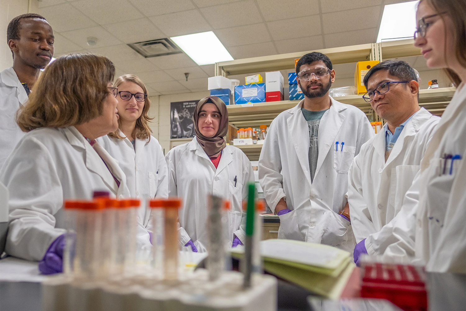 Several scientists in lab coats stand around a table that has lab equipment on it. 