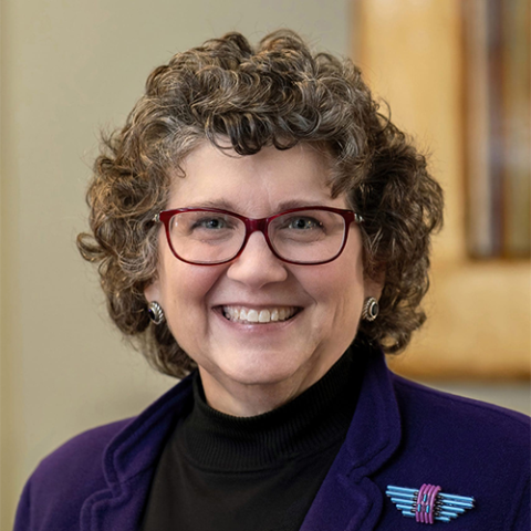 A woman with short, curly brown hair and glasses, wearing a purple jacket, black shirt, and a colorful brooch. 