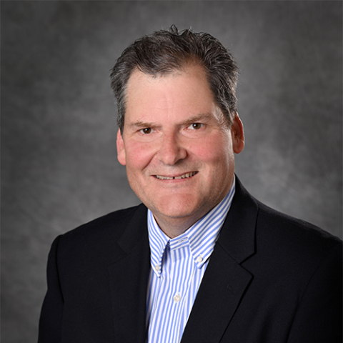 A man with short dark hair, wearing a blue shirt and dark jacket in front of a grey background