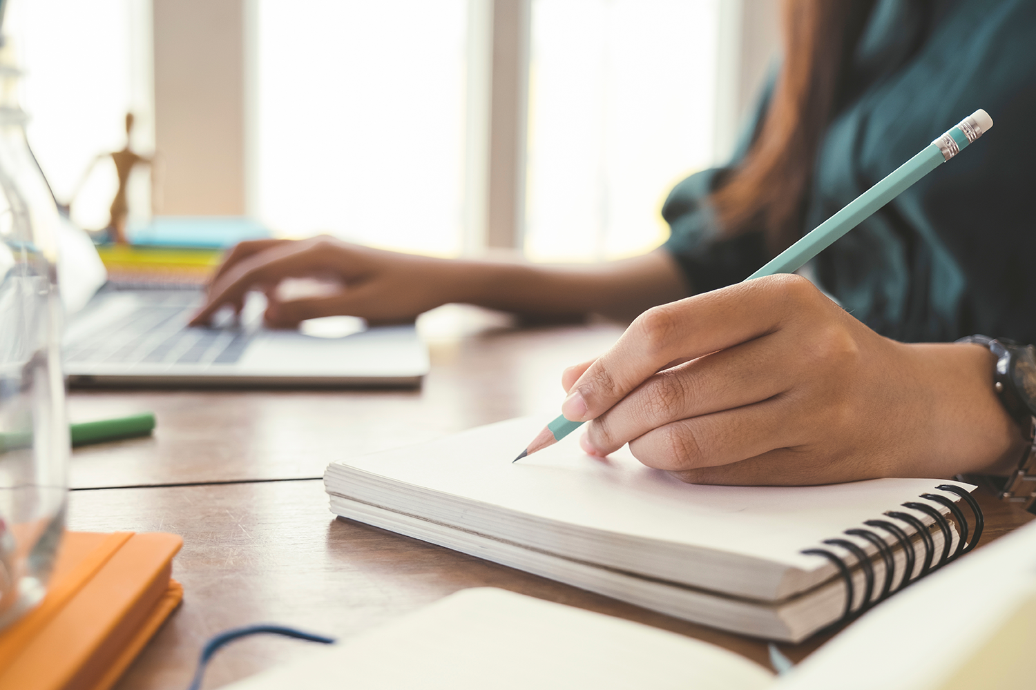 A person using a laptop and writing with a pencil on a notebook