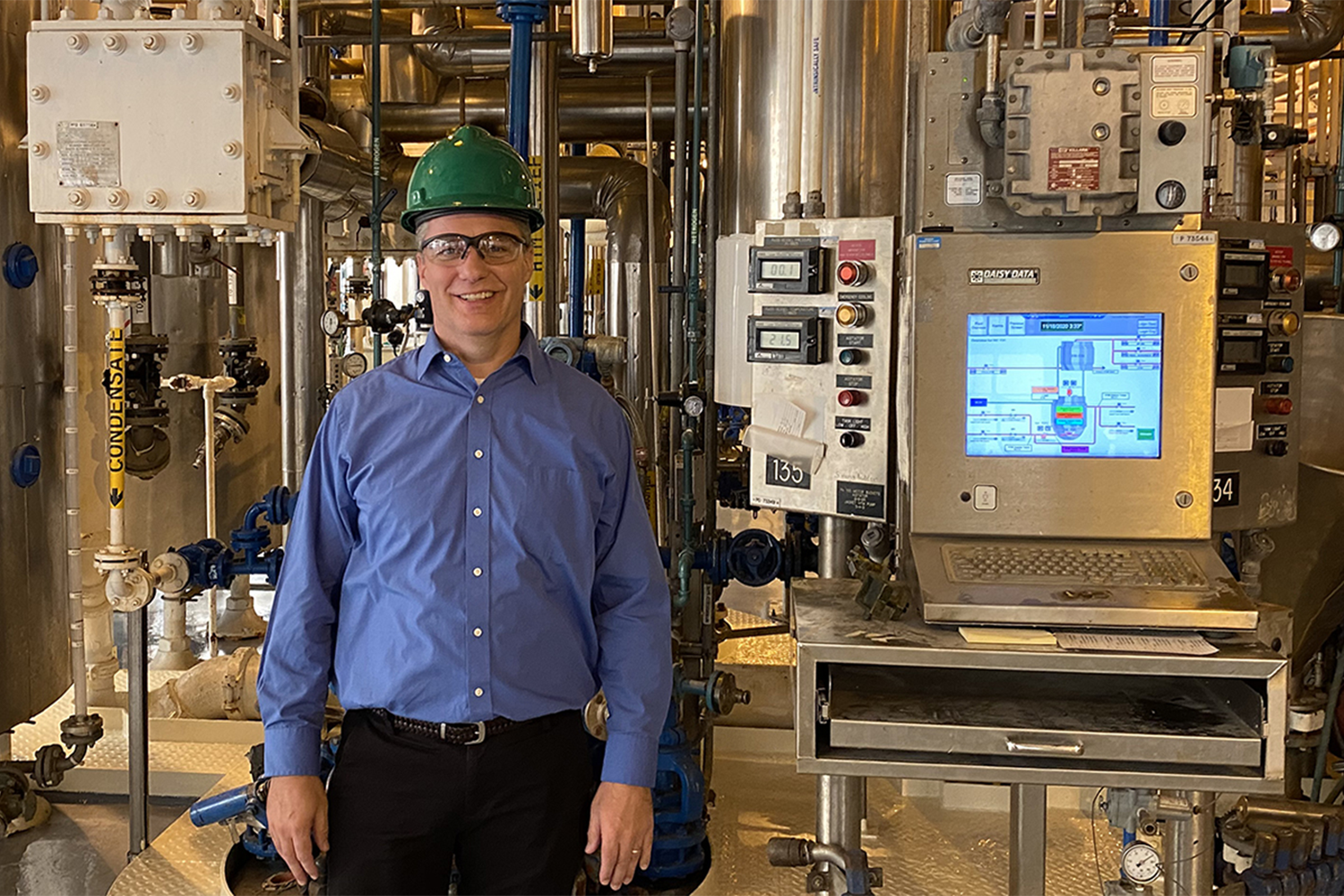 A man wearing a green hard hat and blue shirt, in an industrial manufacturing facility. 