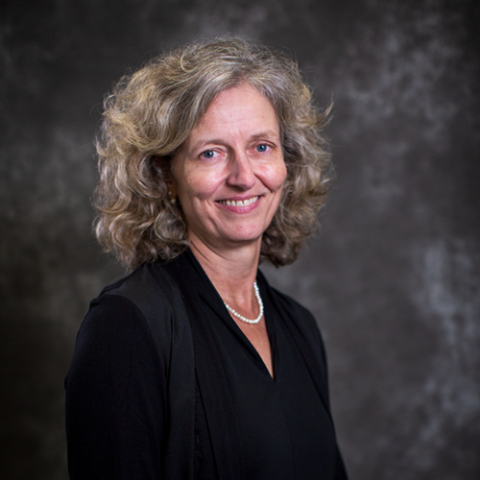 A woman with shoulder length blonde curly hair, and wearing a dark blouse and jacket