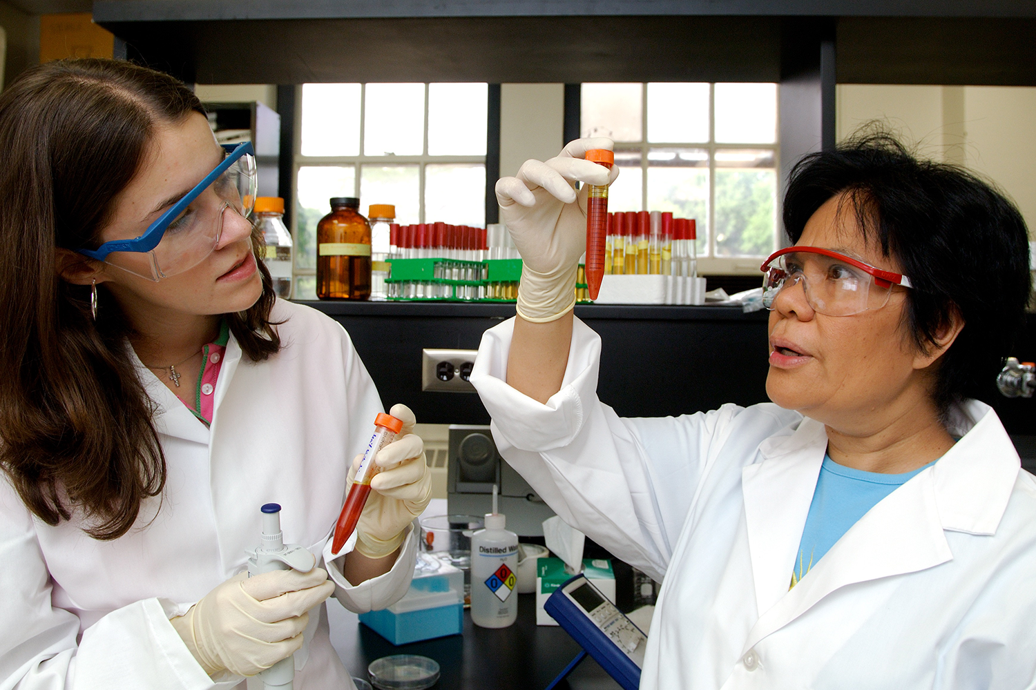 Two scientists, wearing lab coats and safety goggles, holding and looking at test tubes. 