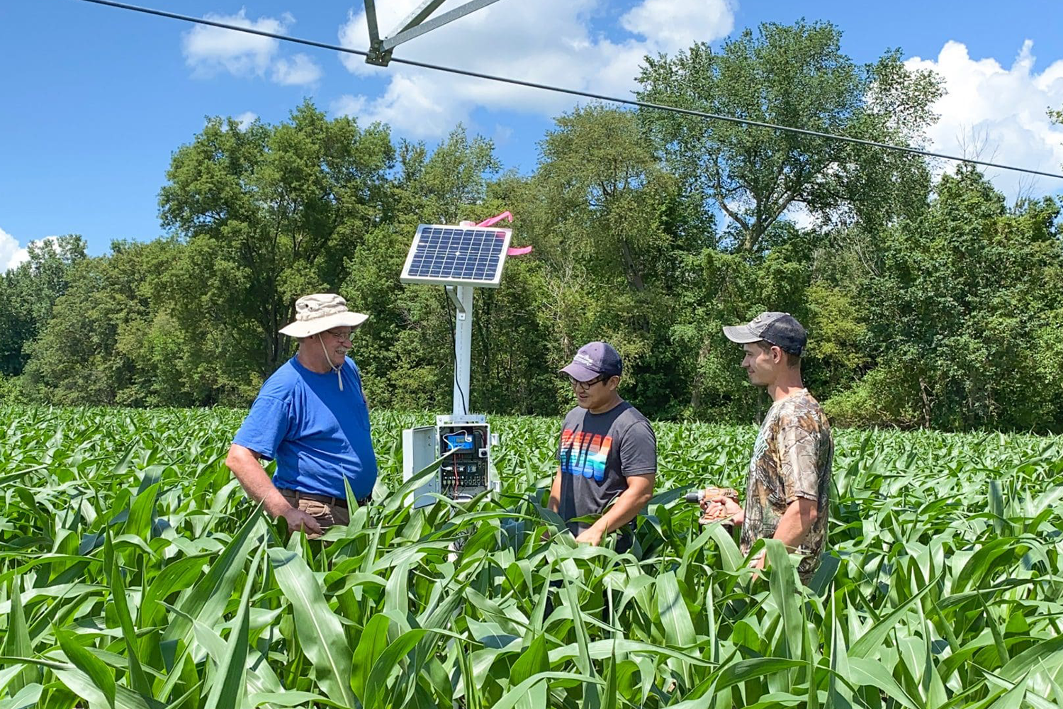 Three people in a field of corn, with a machine that has a solar panel. 